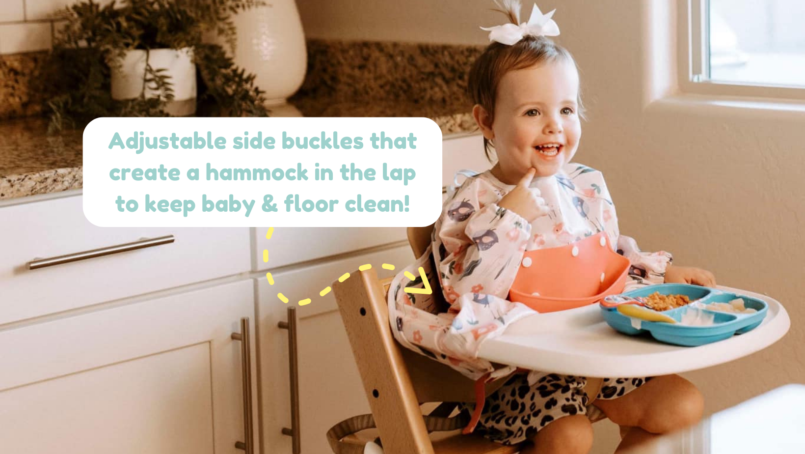 Smiling baby in high chair wearing a Bibvy bib with floral peach design and adjustable side buckles that create a hammock to catch food
