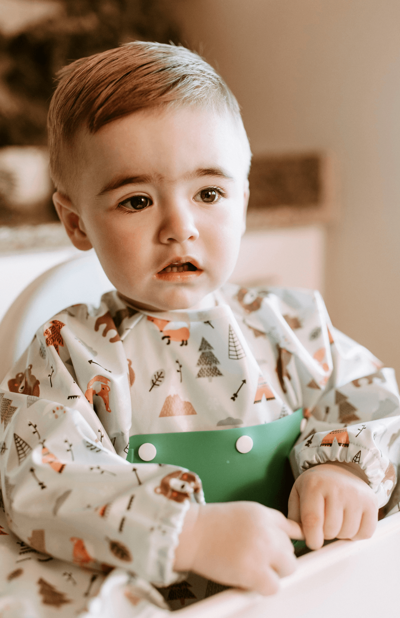 Thoughtful toddler wearing a Bibvy bib in Wilderness Wonder print with a green silicone food catcher, ready for mealtime.