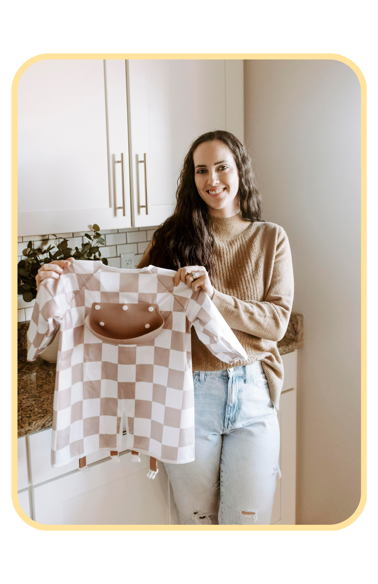 Danielle, the founder of Bibvy, smiling and holding an mauve pink and white checkered bib in a modern kitchen setting.